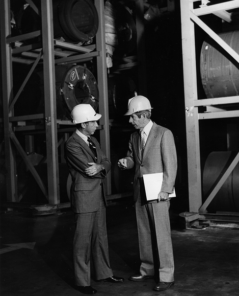 Two executives wearing hard hats talk beside stacked steel coils in a warehouse.