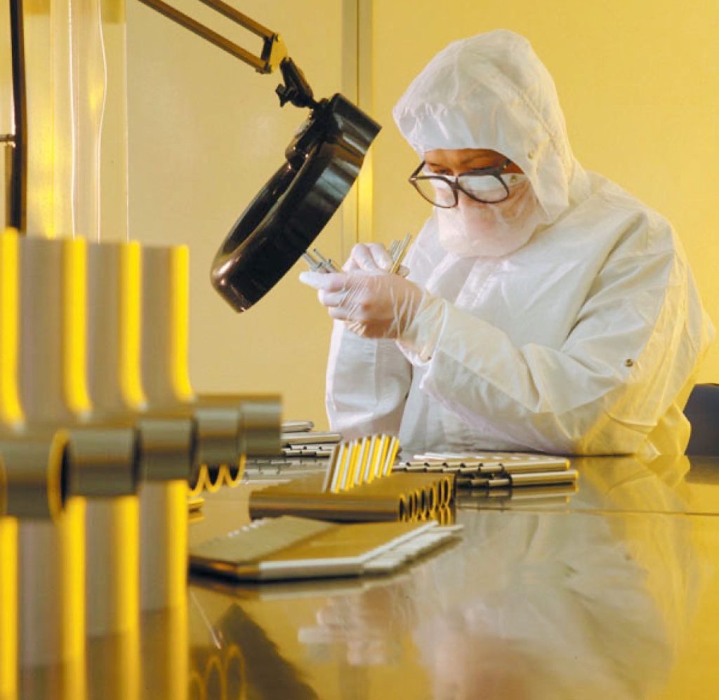 Technician in cleanroom gear inspects precision metal components under magnification.