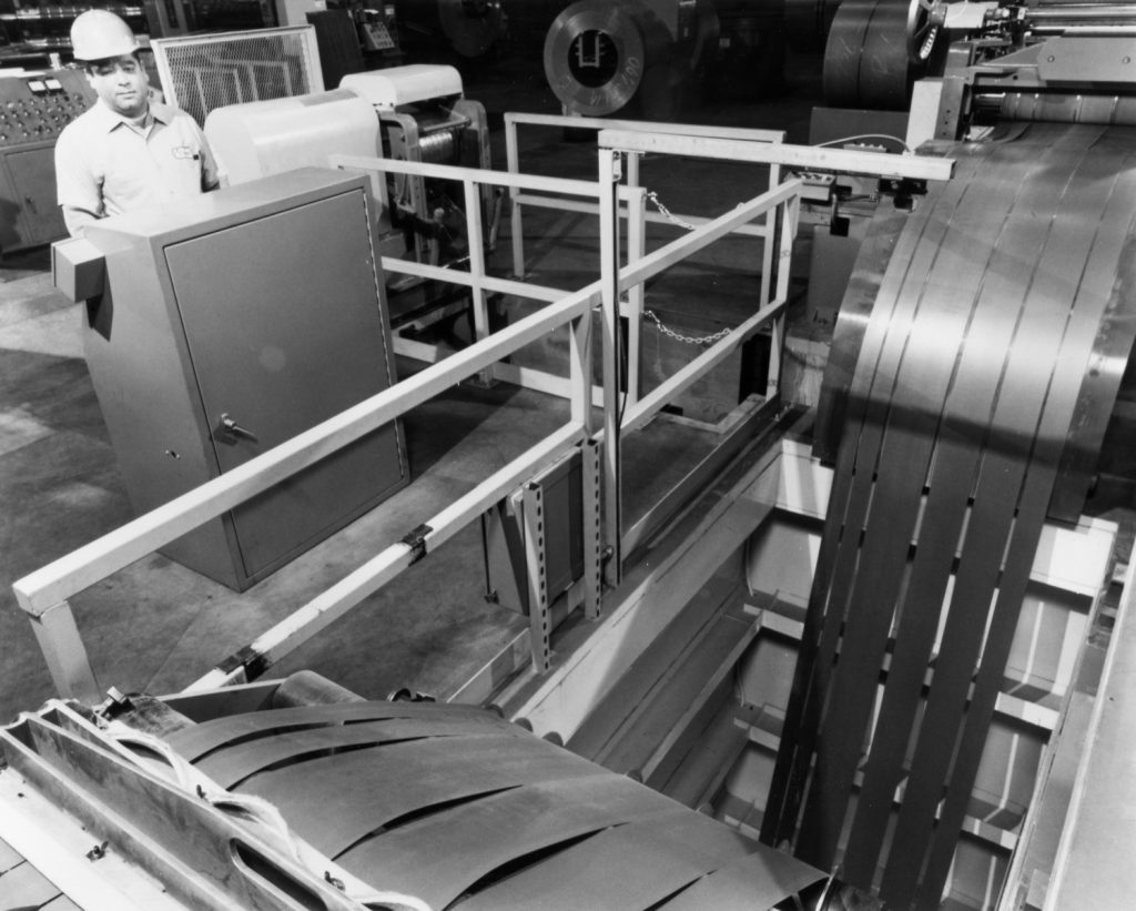Worker monitors a metal slitting machine processing sheet coils in black and white.