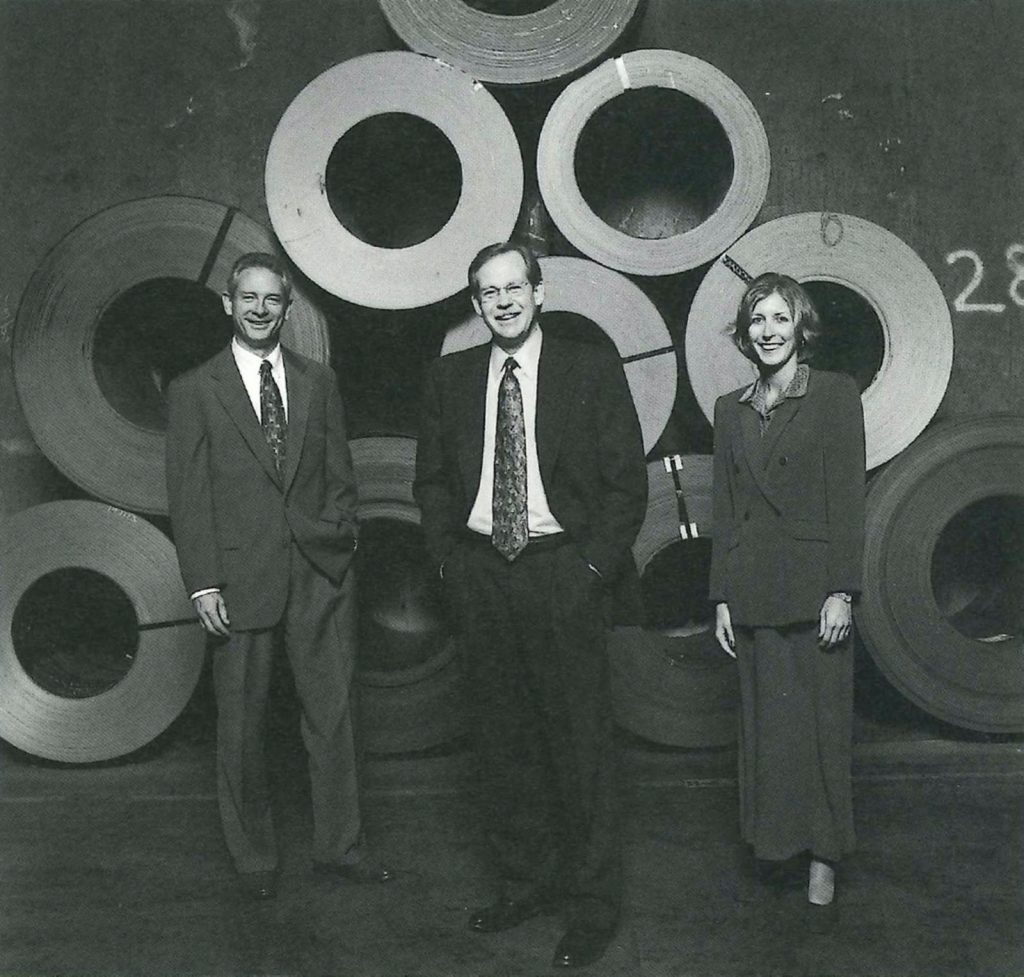 Three executives stand in front of large steel coils in a black-and-white photo.