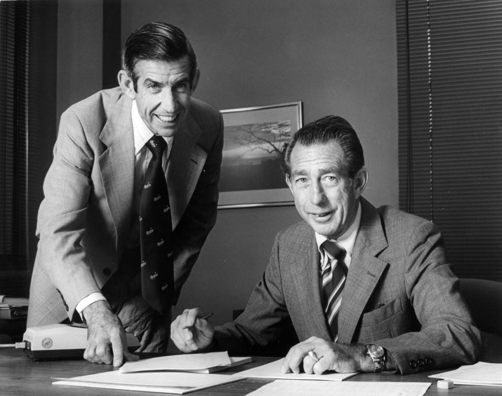 Two Reliance executives pose in an office beside documents on a desk.