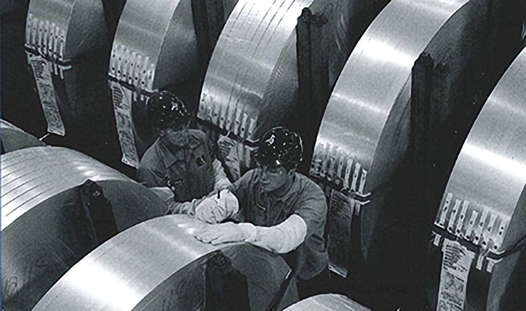 Two workers examine large steel coils in a black-and-white industrial photo.