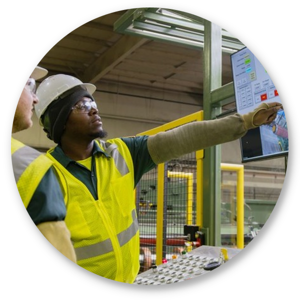 Two workers in safety gear reviewing production data on a large monitor in a manufacturing facility.